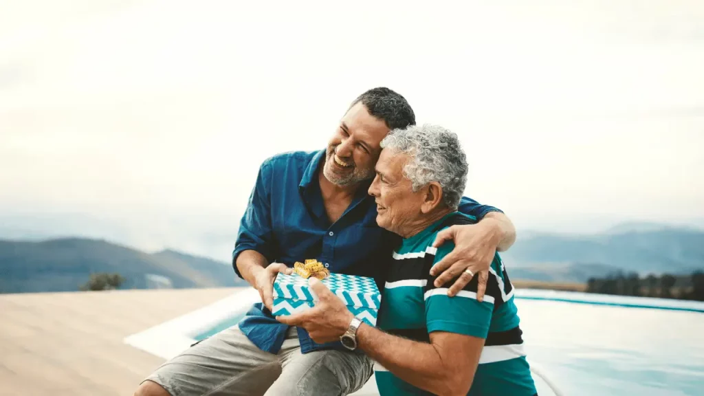 Homem mais novo abraçando e dando um presente para um homem mais velho, em um lugar ao ar livre. 
