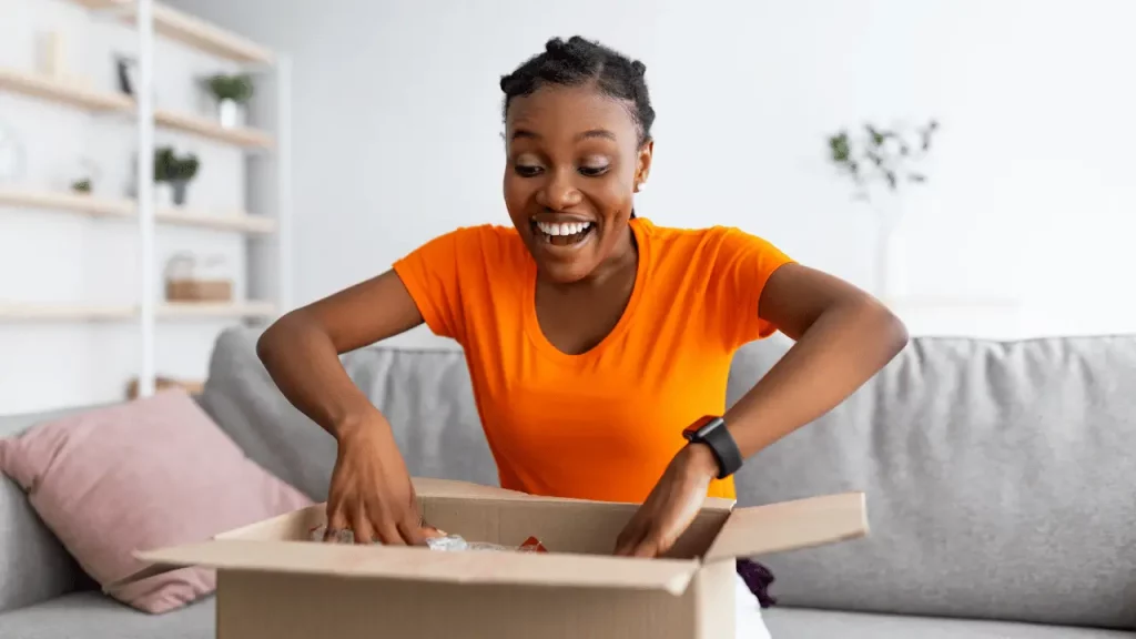 Mulher abrindo uma caixa de encomenda. Ela é negra e veste uma blusa de cor laranja.