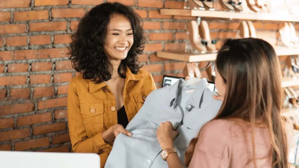 Vendedora mostrando uma camisa social para uma cliente. As duas mulheres são brancas. A vendedoras está usando uma camisa amarela, tem traços orientais e cabelos castanhos. A cliente está de costas, usa uma camisa rosa e cabelo castanho claro. 